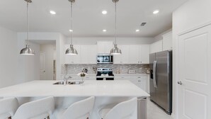 Una cocina moderna con una isla central, electrodomésticos de acero inoxidable, iluminación colgante y armarios blancos.