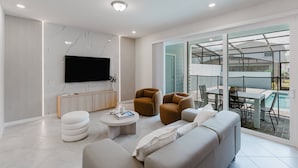 Una sala de estar moderna con un sofá, sillones, una mesa de centro y un televisor. Hay un balcón con una mesa y sillas, y vistas al exterior.