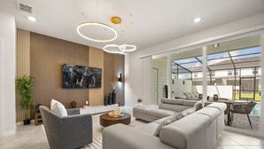 Un salon moderne avec un grand canapé d’angle, une table basse et un téléviseur à écran plat fixé au mur. Une grande fenêtre offre une vue sur l’extérieur.