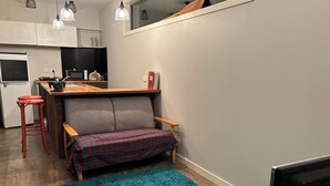 Un espace de vie compact comprenant un coin cuisine, un canapé recouvert d’un plaid, un tapis turquoise et un téléviseur.