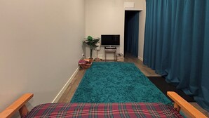 Une petite pièce agréable, avec un tapis vert turquoise, un lit recouvert d’une couverture à carreaux, une télévision, une plante et une fenêtre avec des rideaux.