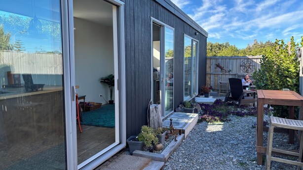 Une maison moderne dotée de grandes portes-fenêtres coulissantes, d’un patio avec une table et des chaises en bois, et d’un jardin agrémenté de diverses plantes.
