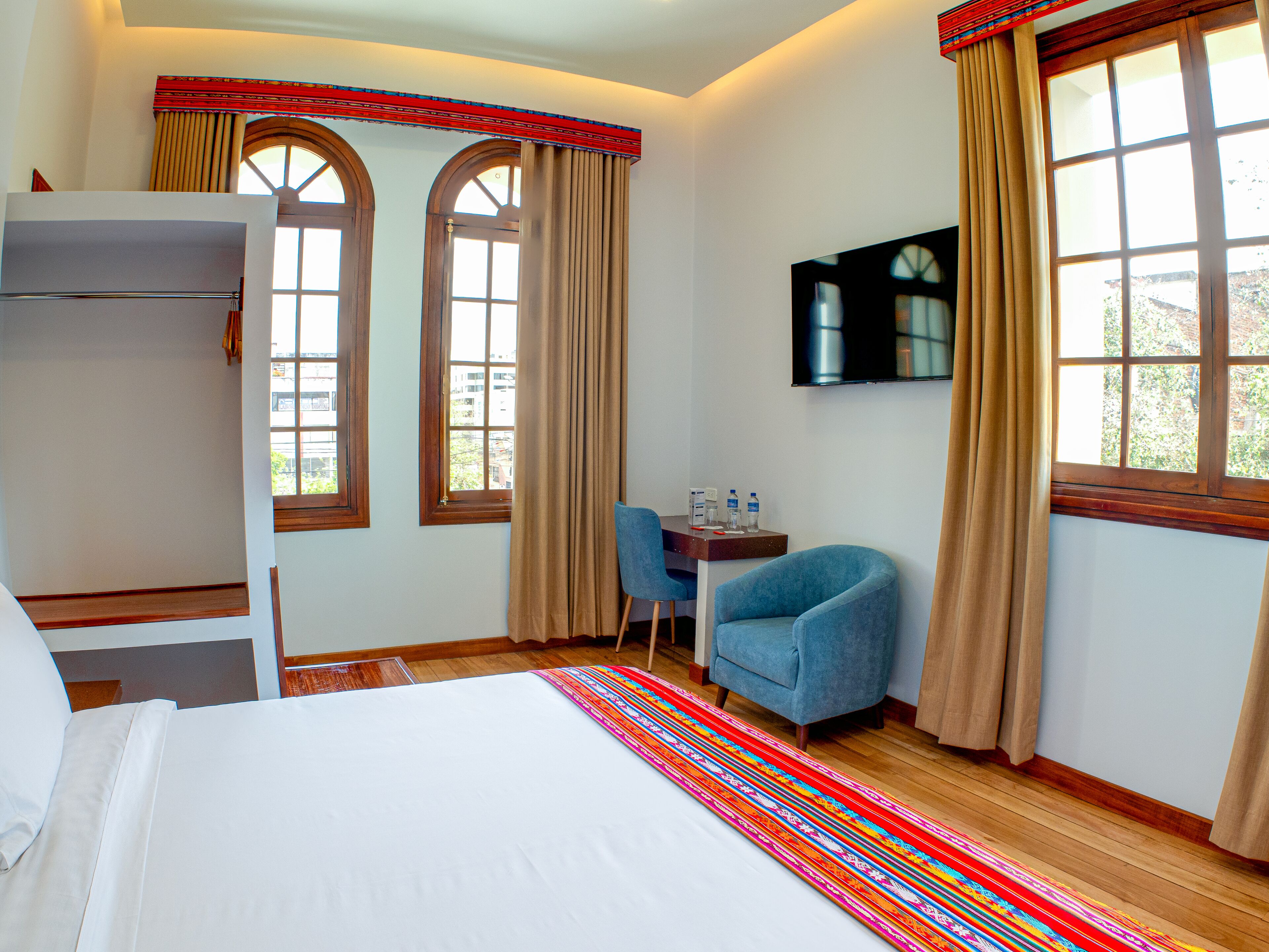 A hotel room with a bed, a desk with a chair, two windows with curtains, and a television on the wall.