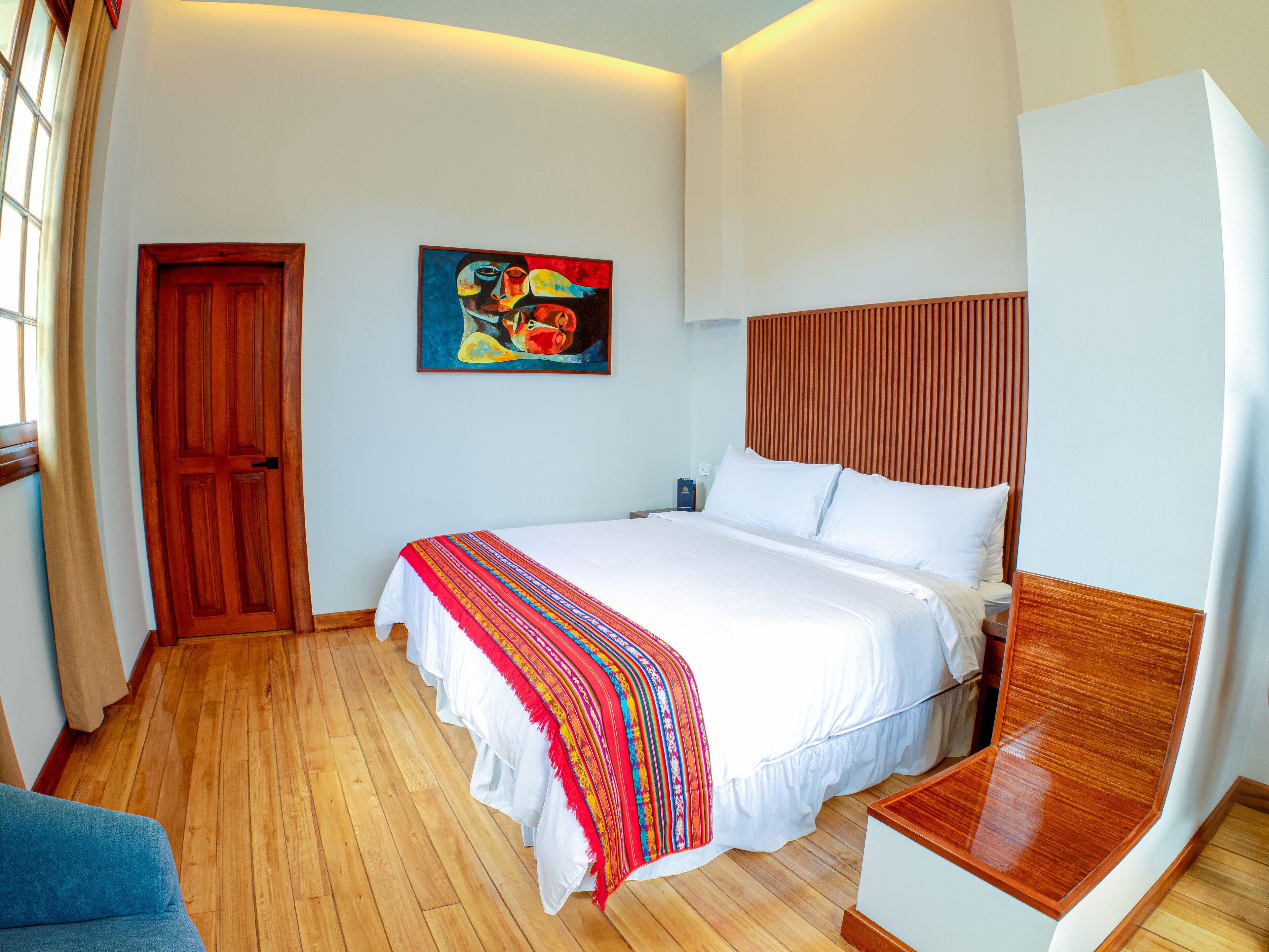 Una habitación de hotel con suelo de madera, una cama con ropa blanca y una manta colorida, un cabecero de madera, un cuadro en la pared y una puerta de madera.