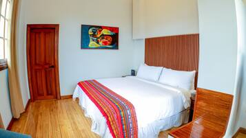 Una habitación de hotel con suelo de madera, una cama con ropa blanca y una manta colorida, un cabecero de madera, un cuadro en la pared y una puerta de madera.