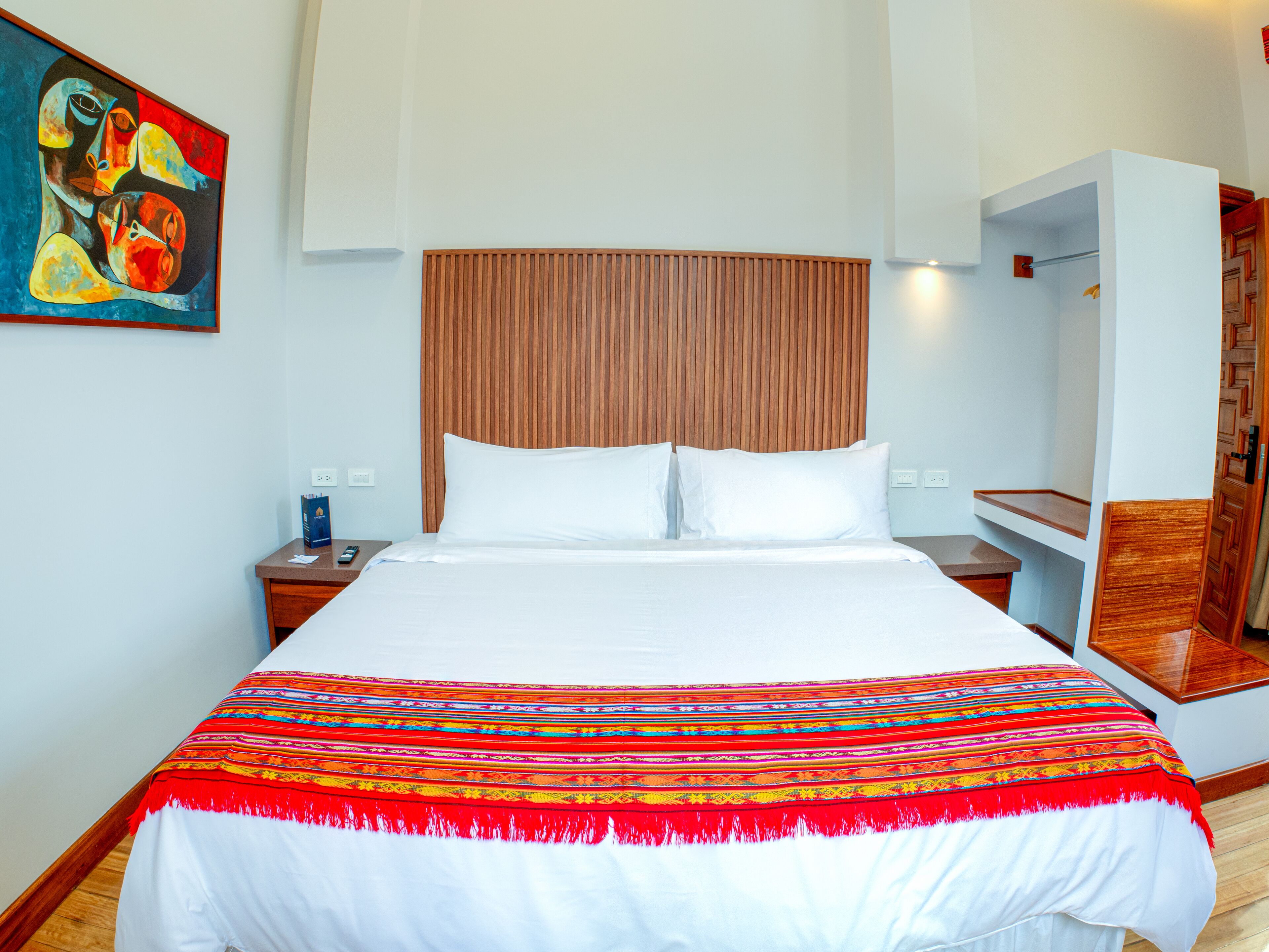 A hotel room with a large bed, a wooden headboard, a colorful blanket, a nightstand, a painting on the wall, and a wooden wardrobe.