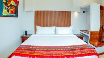A hotel room with a large bed, a wooden headboard, a colorful blanket, a nightstand, a painting on the wall, and a wooden wardrobe.