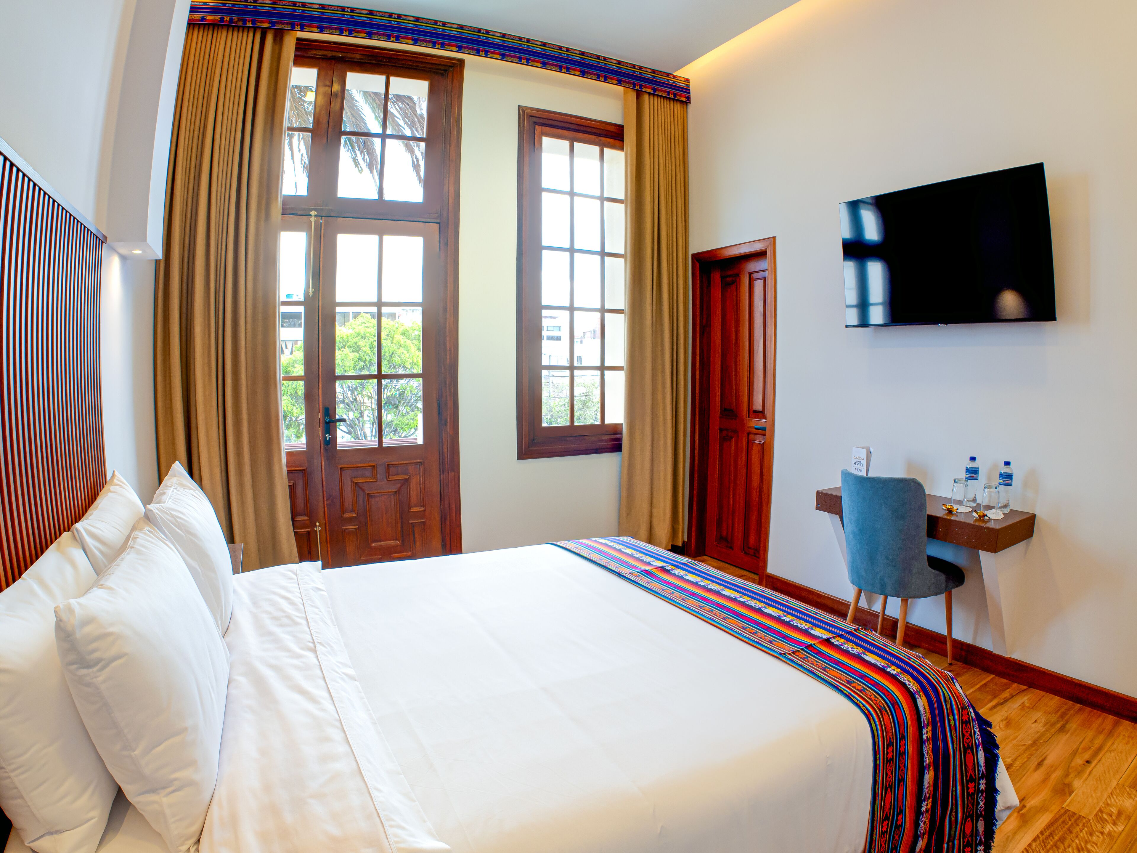 A hotel room with a large bed, two windows with curtains, a TV on the wall, and a small desk with a chair.