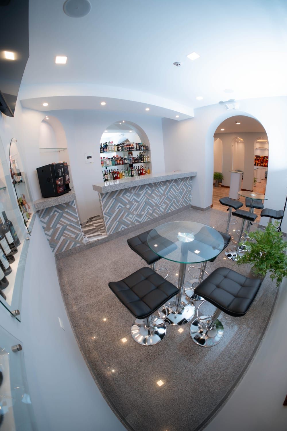 A modern bar area with a glass-top table and black cushioned stools, a bar counter with a variety of bottles, and a geometric patterned wall.