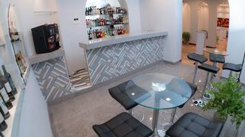 A modern bar area with a glass-top table and black cushioned stools, a bar counter with a variety of bottles, and a geometric patterned wall.