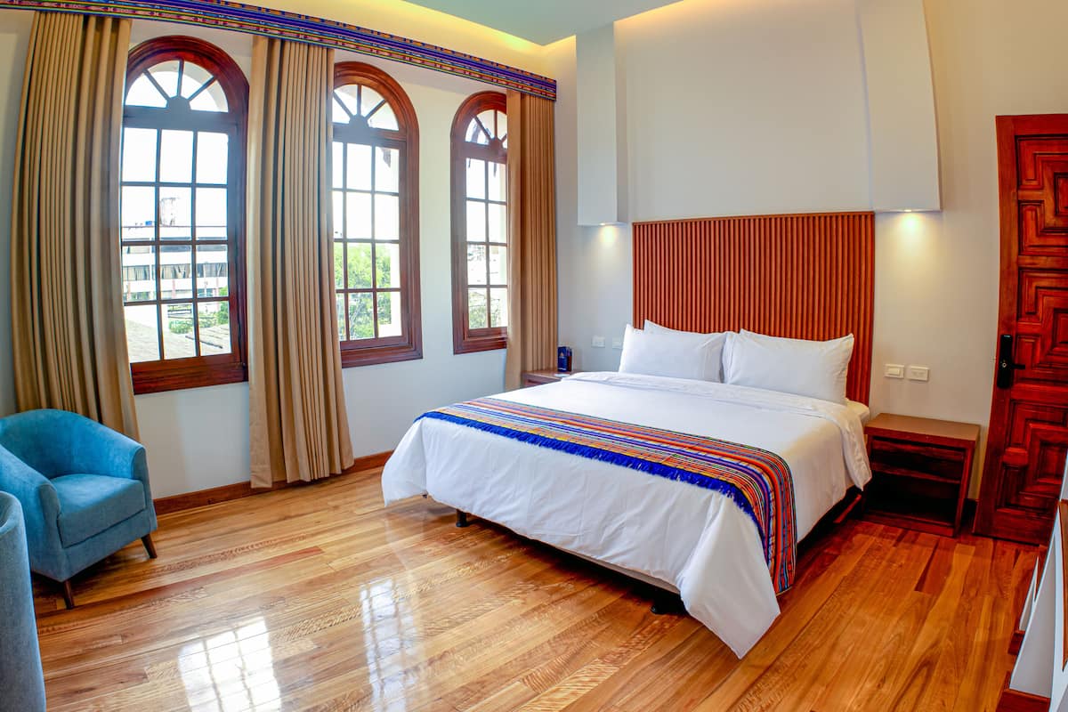 Een hotelkamer met een groot bed, houten vloeren, een blauwe fauteuil en een raam met gordijnen.