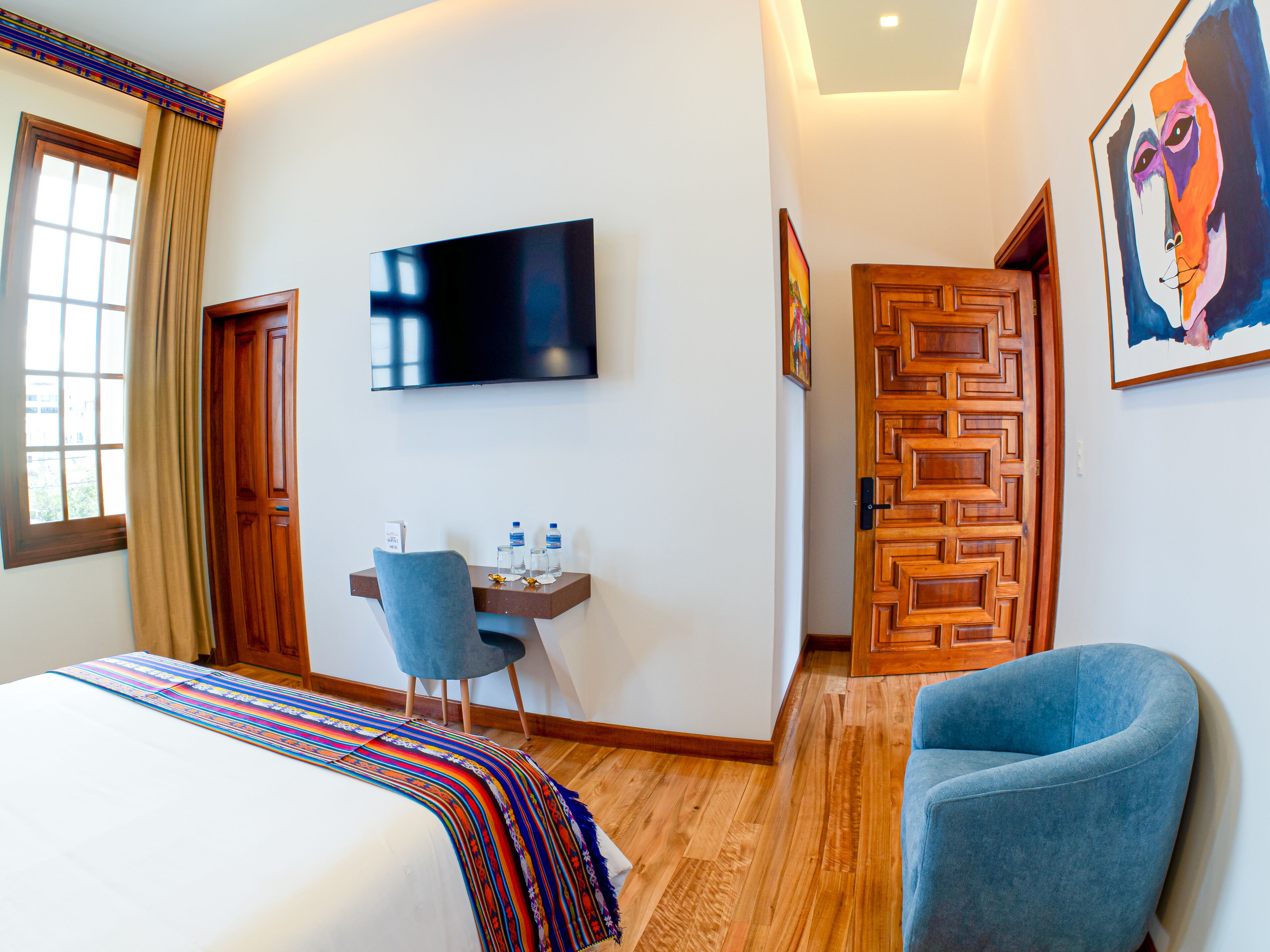A hotel room with a bed, a desk with a chair, a TV, and a door with a geometric pattern.