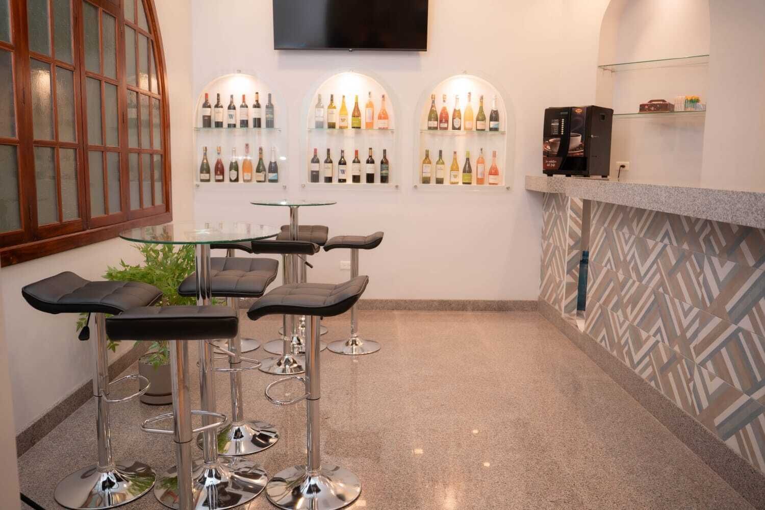 A modern bar area with glass tables and black bar stools, a TV mounted on the wall, and shelves with bottles of beverages.