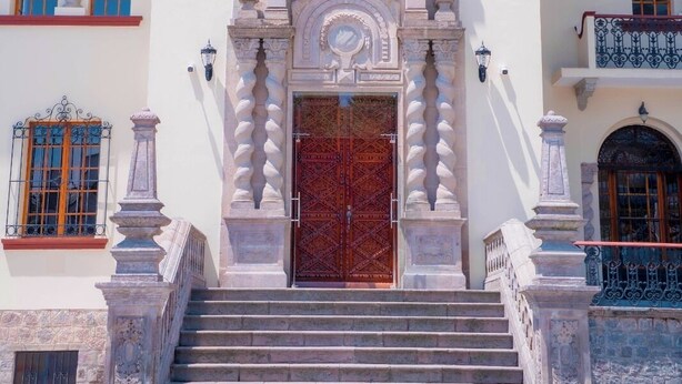 Un edificio de dos pisos con una entrada central que presenta una amplia puerta de madera, flanqueada por columnas de piedra y arcos decorativos.