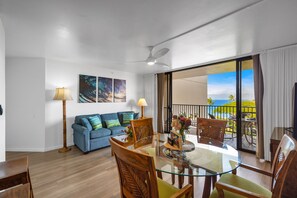 Ein modernes Wohnzimmer mit einem Glastisch, Holzstühlen, einem blauen Sofa und einem Balkon mit Meerblick.