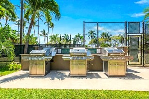 Un espace barbecue extérieur doté de trois grils, entouré de palmiers et délimité par une clôture.