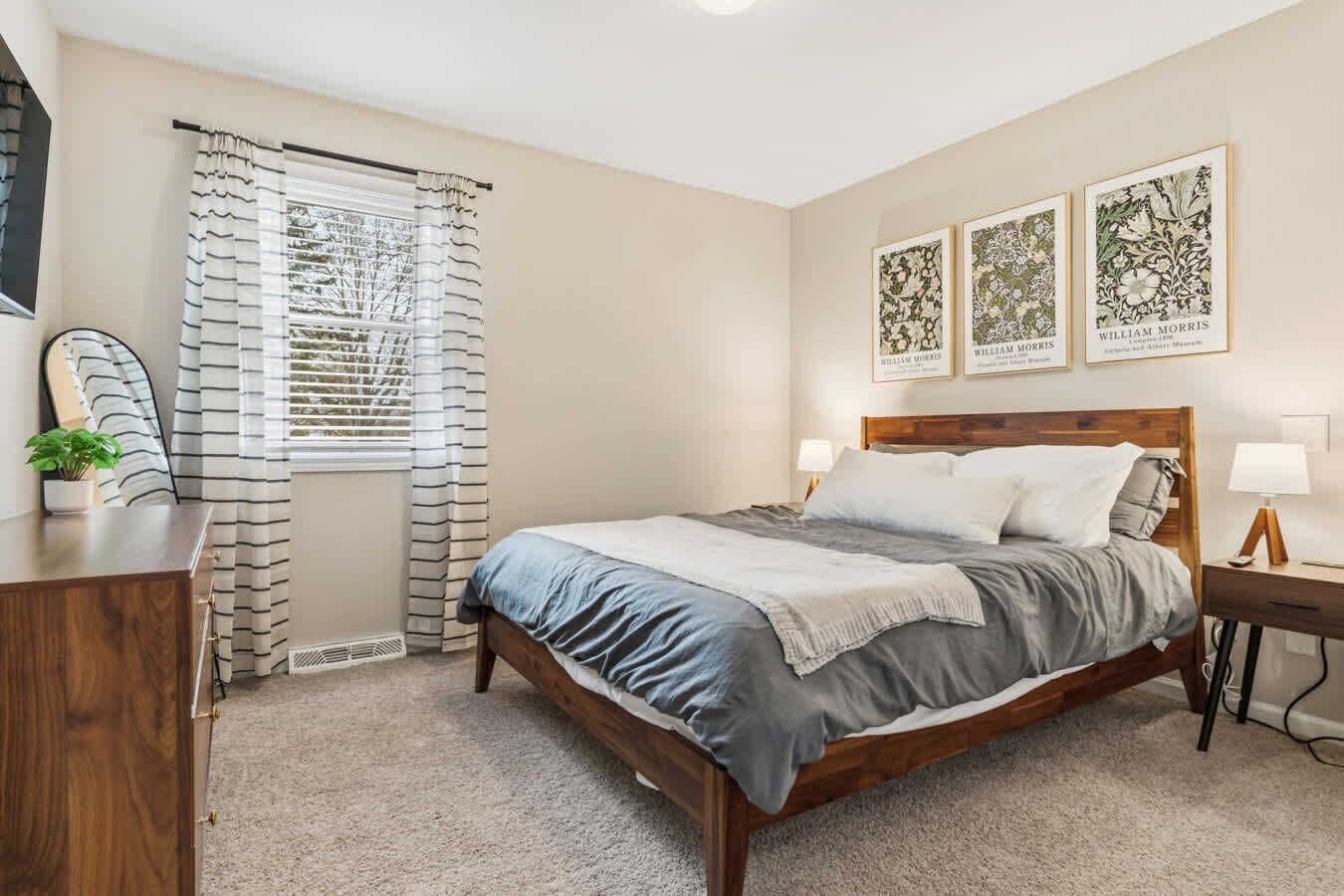 A bedroom with a wooden bed, a desk, a chair, a window with striped curtains, and three framed botanical prints on the wall.