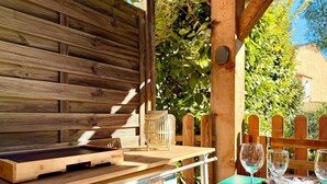 An outdoor dining area with wooden tables and chairs, surrounded by greenery and a wooden fence. - Le Cocon du Mas, 4 guests, Pool in Calvisson (Calvisson)