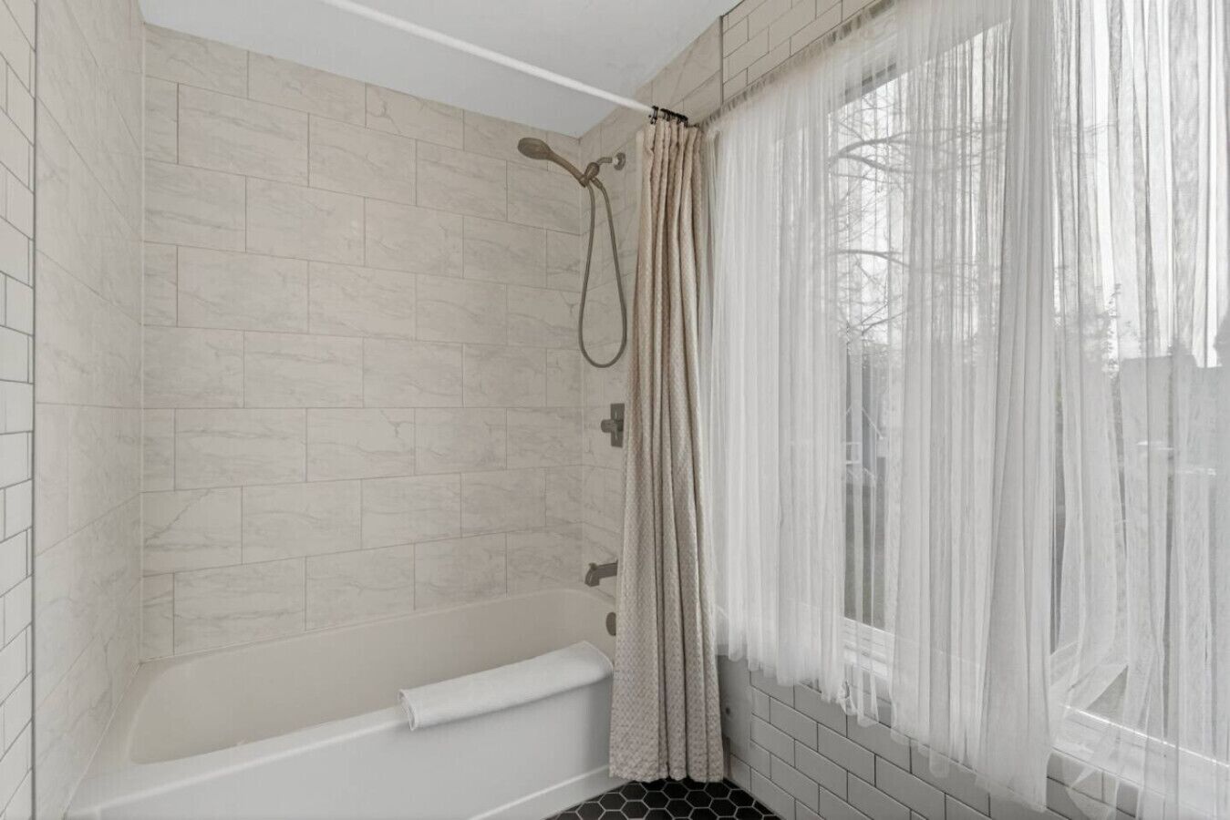 Ein Badezimmer mit Dusche, Badewanne und einem Fenster mit durchsichtigen Vorhängen.