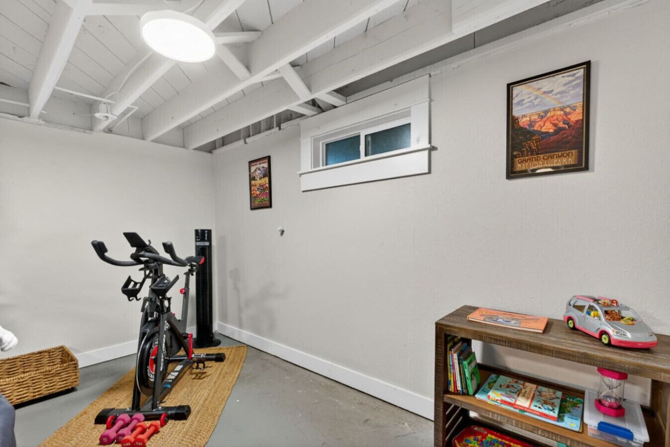A home gym with exercise equipment, a window, and a bookshelf with children's books and toys.
