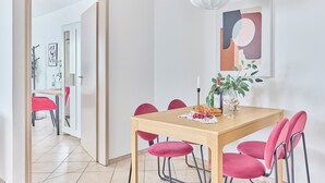 Ruokailutilassa on puinen pöytä, kuusi vaaleanpunaista tuolia, valkoinen riippuvalaisin ja seinällä taulu.