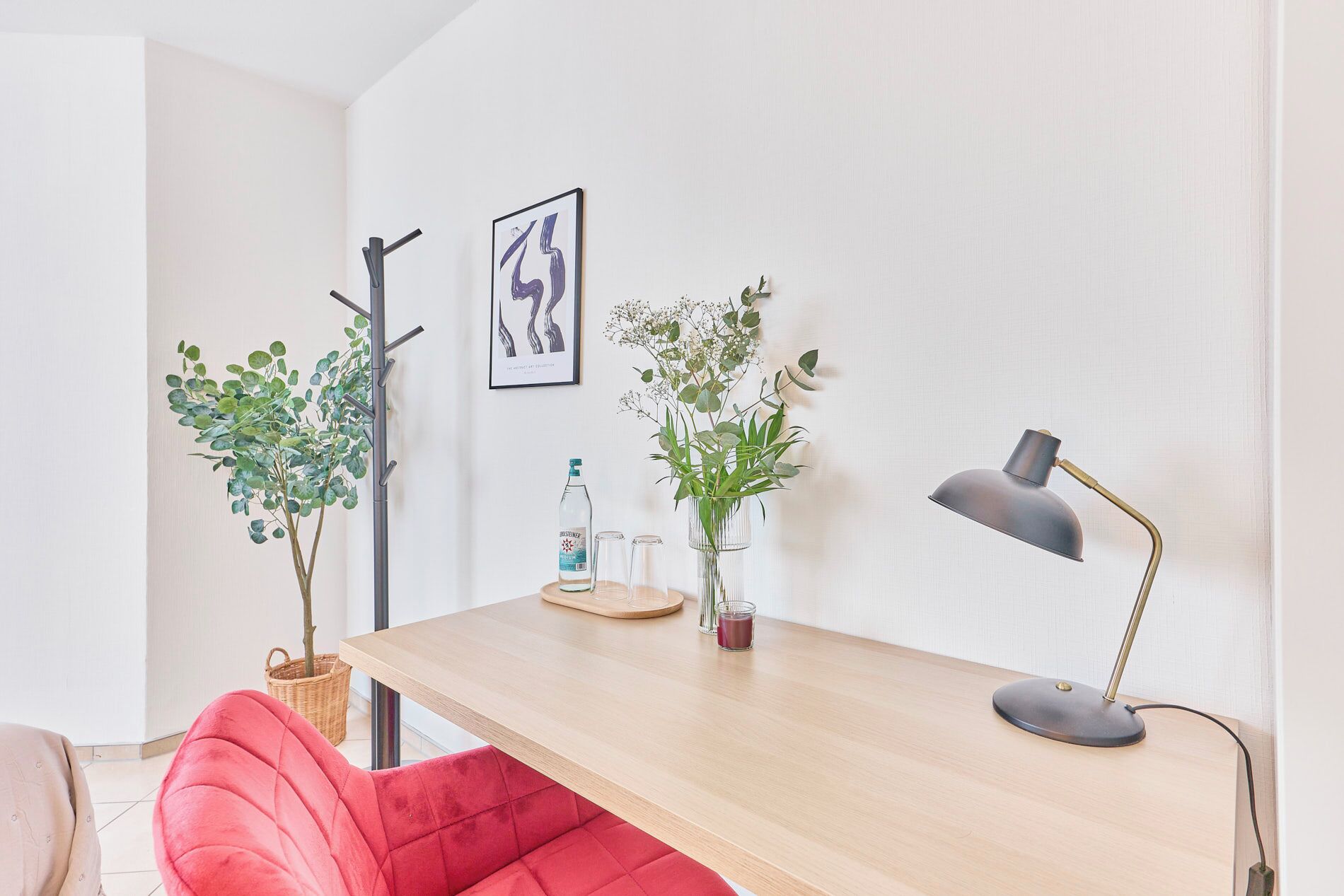 Una habitación moderna con un escritorio de madera, una silla roja, dos plantas en macetas, una obra de arte enmarcada y una lámpara de escritorio.