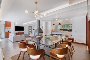 Een ruime woonkamer met een grote eettafel met glazen blad, houten stoelen, een moderne haard en een keuken op de achtergrond.