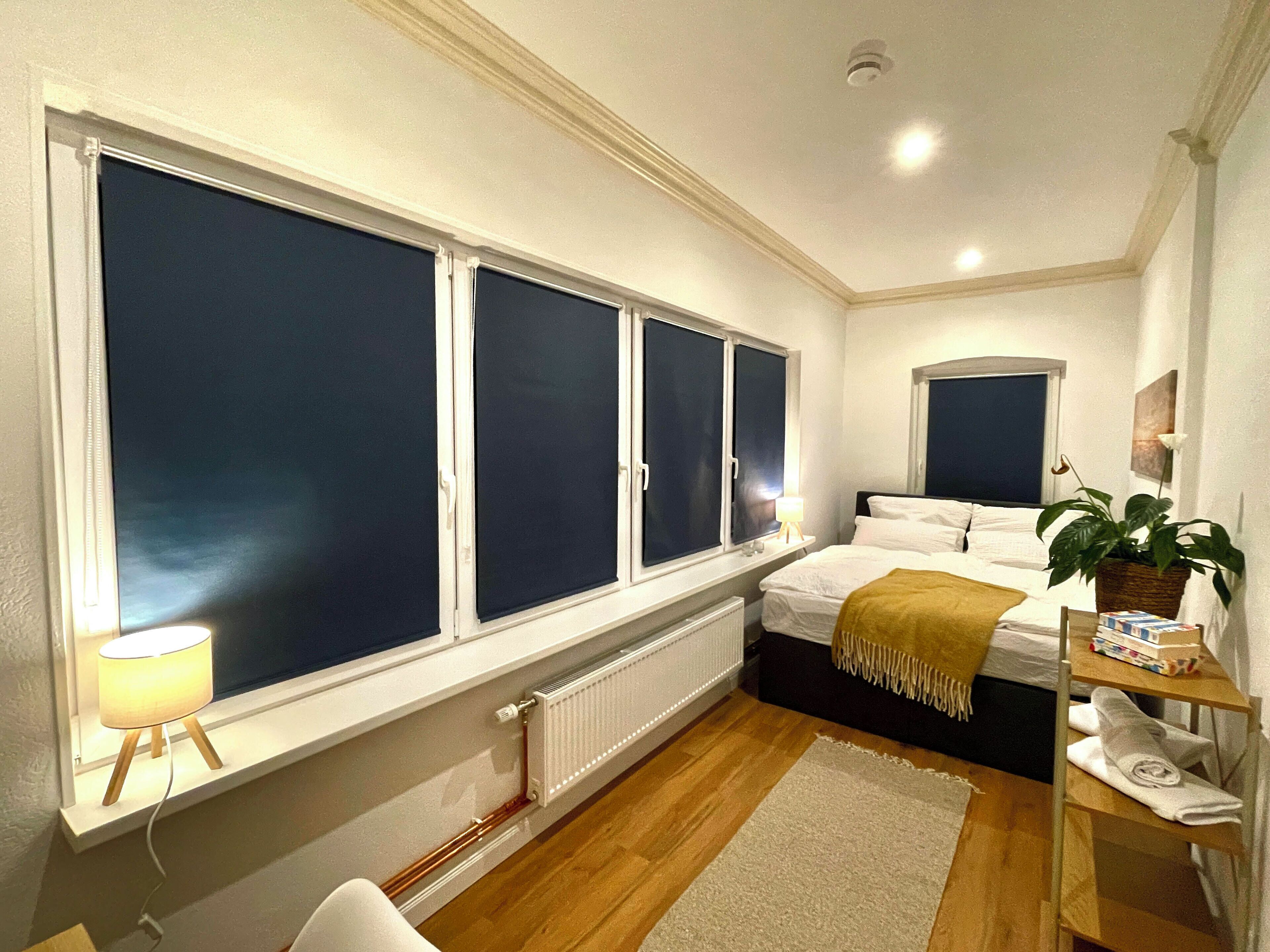 Une chambre avec une grande fenêtre, un lit, une table de chevet avec une lampe, un radiateur et une étagère avec des serviettes.