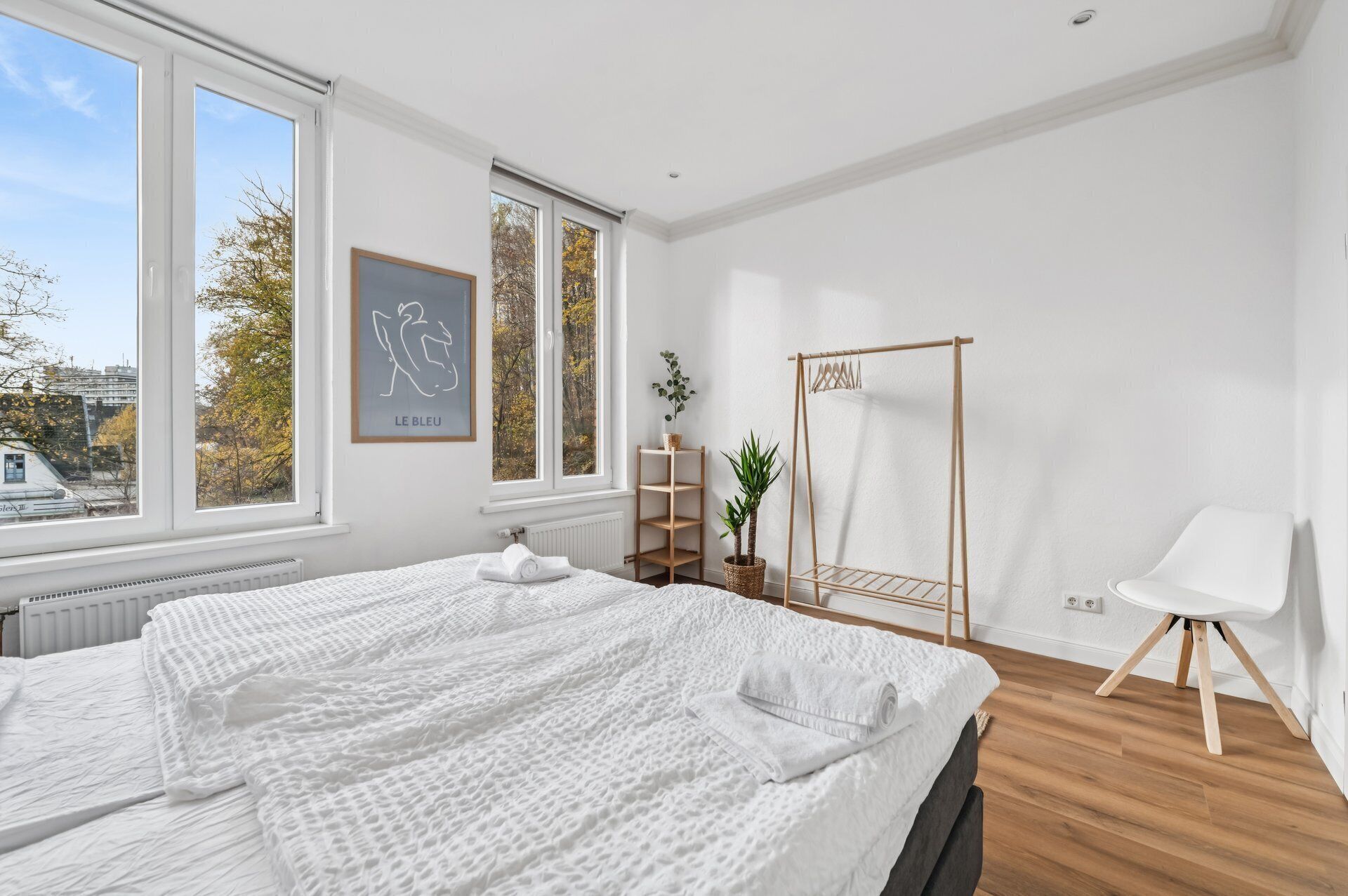 Een moderne slaapkamer met een groot bed, een houten kledingrek, een witte stoel en uitzicht op herfstbomen door het raam.