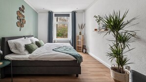 Une chambre à coucher comprenant un lit, une table de chevet, une fenêtre avec des rideaux, une plante sur la table de chevet et une étagère fixée au mur.