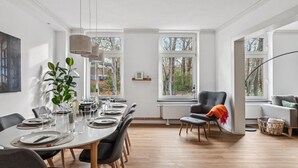 Un coin repas avec une table ronde dressée pour six personnes, entourée de chaises. De grandes fenêtres offrent une vue sur les arbres et un bâtiment à l’extérieur.