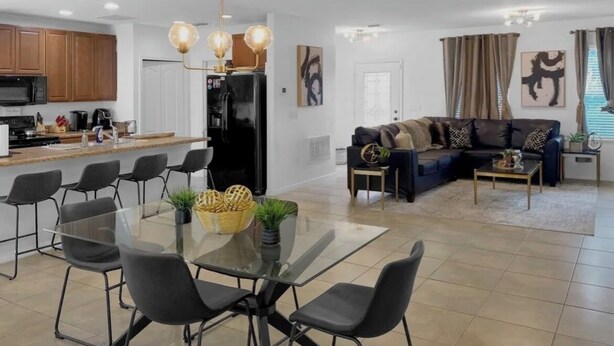 A modern kitchen with wooden cabinets, a black refrigerator, and a dining area with a glass table and black chairs. - Charming 3-bedroom house with AC in peaceful neighborhood, minutes from Disney! (Dundee)