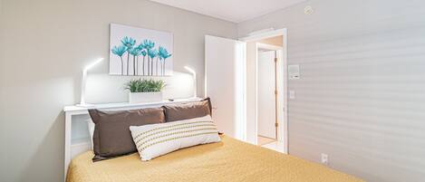 A bedroom with a bed, a nightstand, a wall-mounted artwork, and a door leading to another room.
