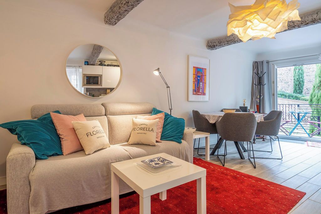 A living room with a sofa, a coffee table, a dining area with chairs, a round mirror, a lamp, and a painting on the wall.
