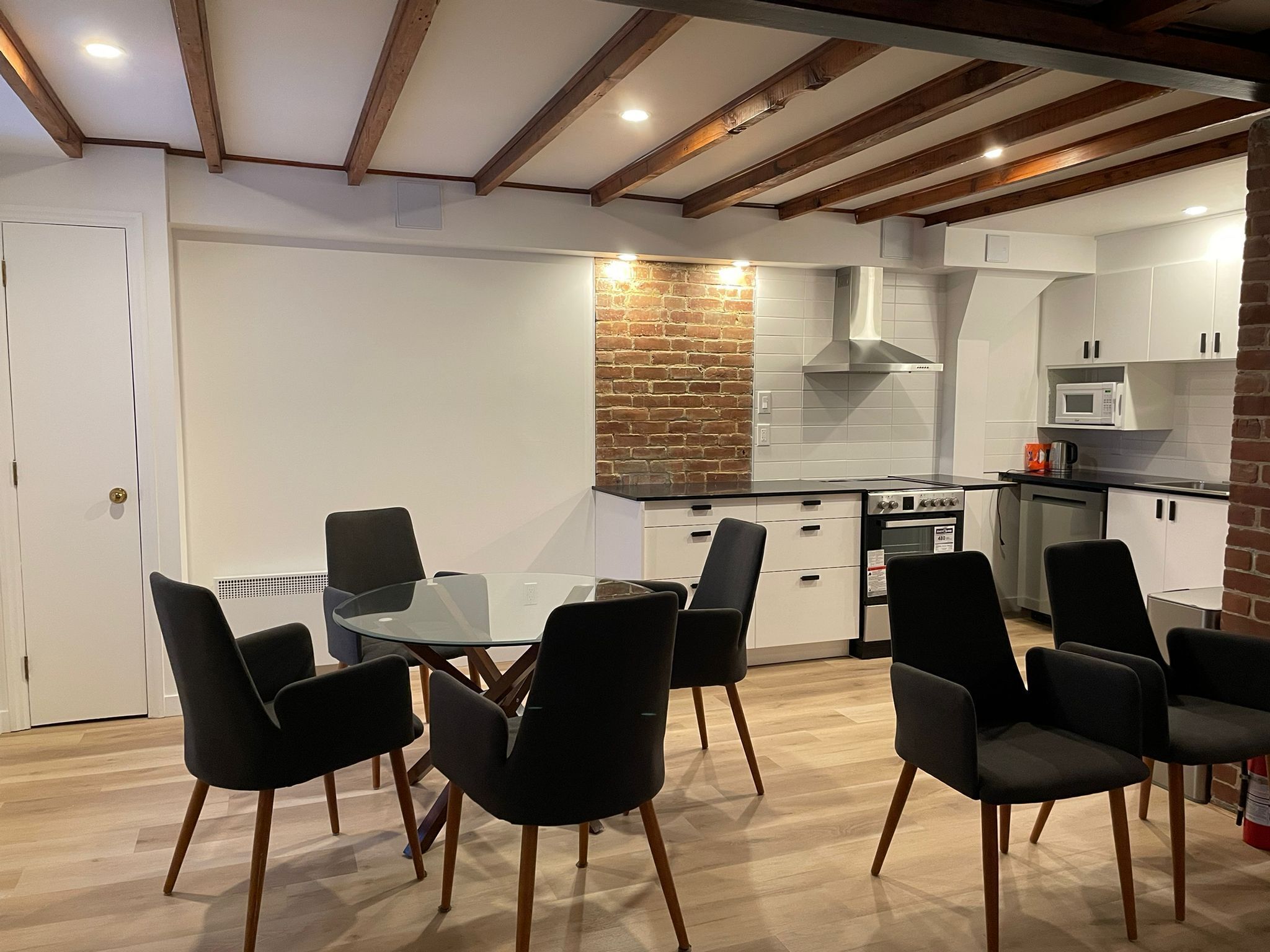 Une cuisine moderne avec des meubles blancs, un mur d’accent en briques et un coin repas avec des chaises noires et une table en verre.