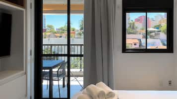 A hotel room with a bed, a window with a city view, a balcony with a table and chairs, and a television on the wall.