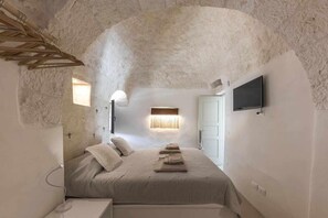 Une chambre avec un mur en pierre, un lit avec des oreillers, une table de chevet, une lampe et un téléviseur fixé au mur.
