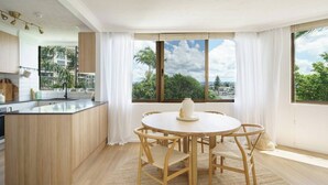 A modern kitchen with a central island, a dining area with a round table and chairs, and large windows offering a view of greenery and buildings. - Palms on Gold Riverfront Haven by Gold Coast Beach (Surfers Paradise)