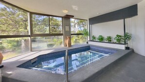 A modern indoor swimming pool with clear glass walls, a tiled floor, and a variety of potted plants. - Palms on Gold Riverfront Haven by Gold Coast Beach (Surfers Paradise)