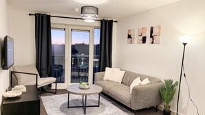 Una sala moderna con un sofá gris, una mesa de centro redonda, un mueble de TV negro y un ventanal grande con cortinas.