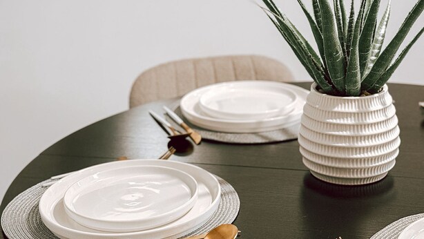 Una mesa de comedor dispuesta con platos blancos, cubiertos dorados y una maceta blanca con textura y una planta verde.