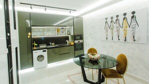 A modern kitchen with a built-in washer and dryer, a glass dining table, and a wall-mounted art piece. - Premium One Bedroom Apartment at KASS (Accra)