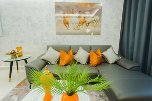 A modern living room with a grey sofa, orange and grey cushions, a glass coffee table, and a framed artwork of horses on the wall. - Premium One Bedroom Apartment at KASS (Accra)