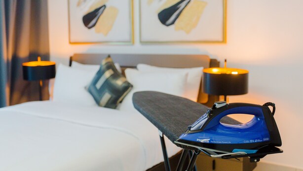 A neatly made bed with pillows, a bedside table with a lamp, and an ironing board with an iron. - Premium One Bedroom Apartment at KASS (Accra)