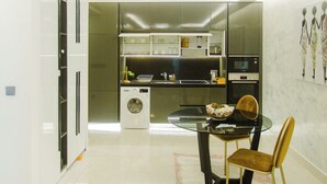 A modern kitchen with a built-in oven, a washing machine, and a dining area with a glass table and chairs. - Premium One Bedroom Apartment at KASS (Accra)