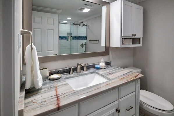 Une salle de bain avec un lavabo en marbre, des meubles blancs, un grand miroir et des toilettes.