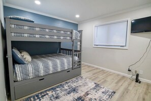 Ein Etagenbett mit grauem Rahmen und gestreifter Bettwäsche, eine blaue Akzentwand, ein Fenster mit Jalousien und ein an der Wand befestigter Fernseher.
