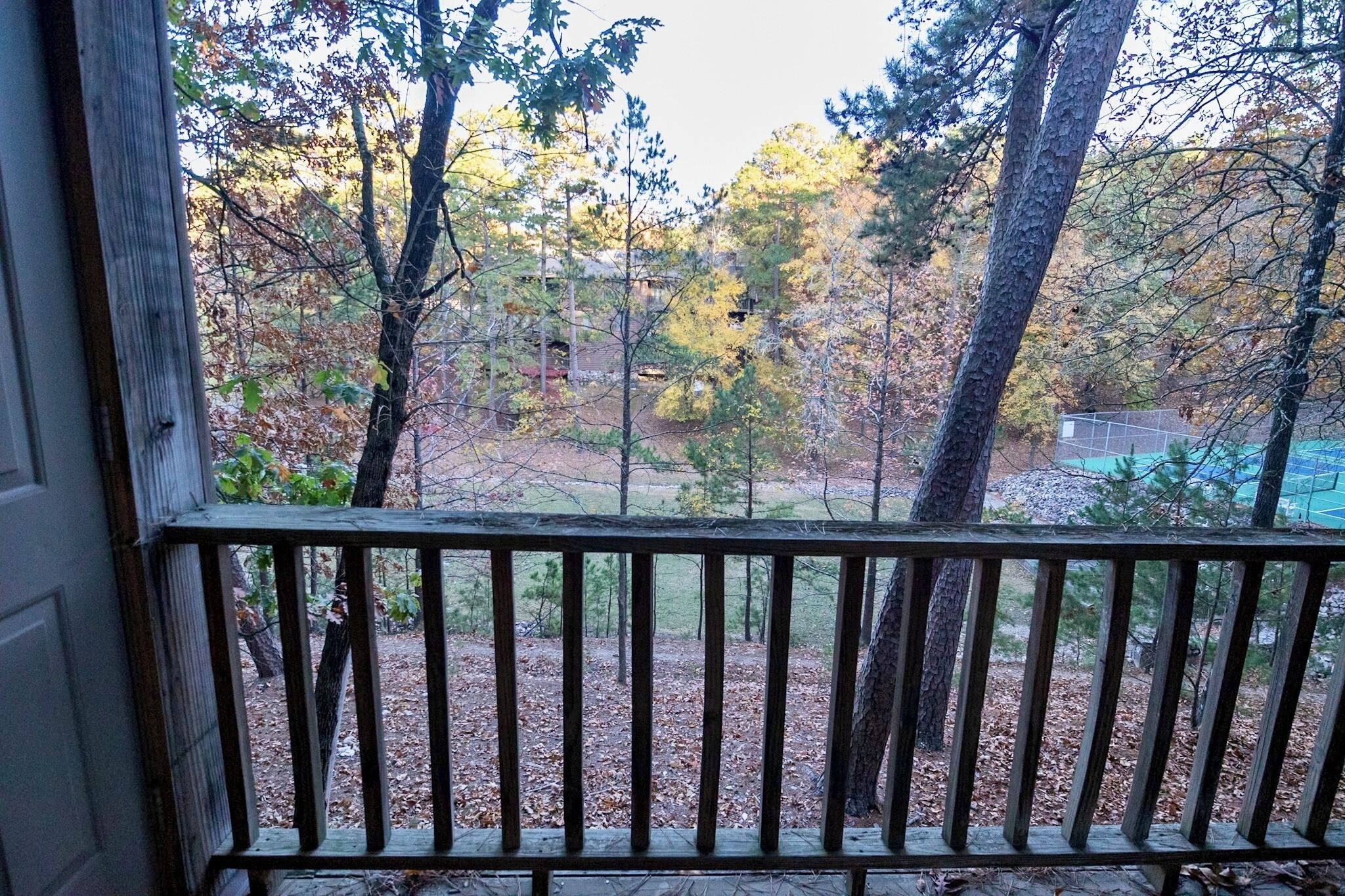 A view from a balcony with a wooden railing, overlooking a wooded area with trees and a grassy field.