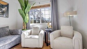 Un salon avec un canapé gris, deux fauteuils blancs, une plante en pot, une fenêtre avec des rideaux et une lampe.