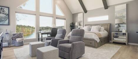 Une chambre spacieuse avec un grand lit, deux fauteuils gris, une petite table et une vue sur un lac, visible à travers de grandes fenêtres.